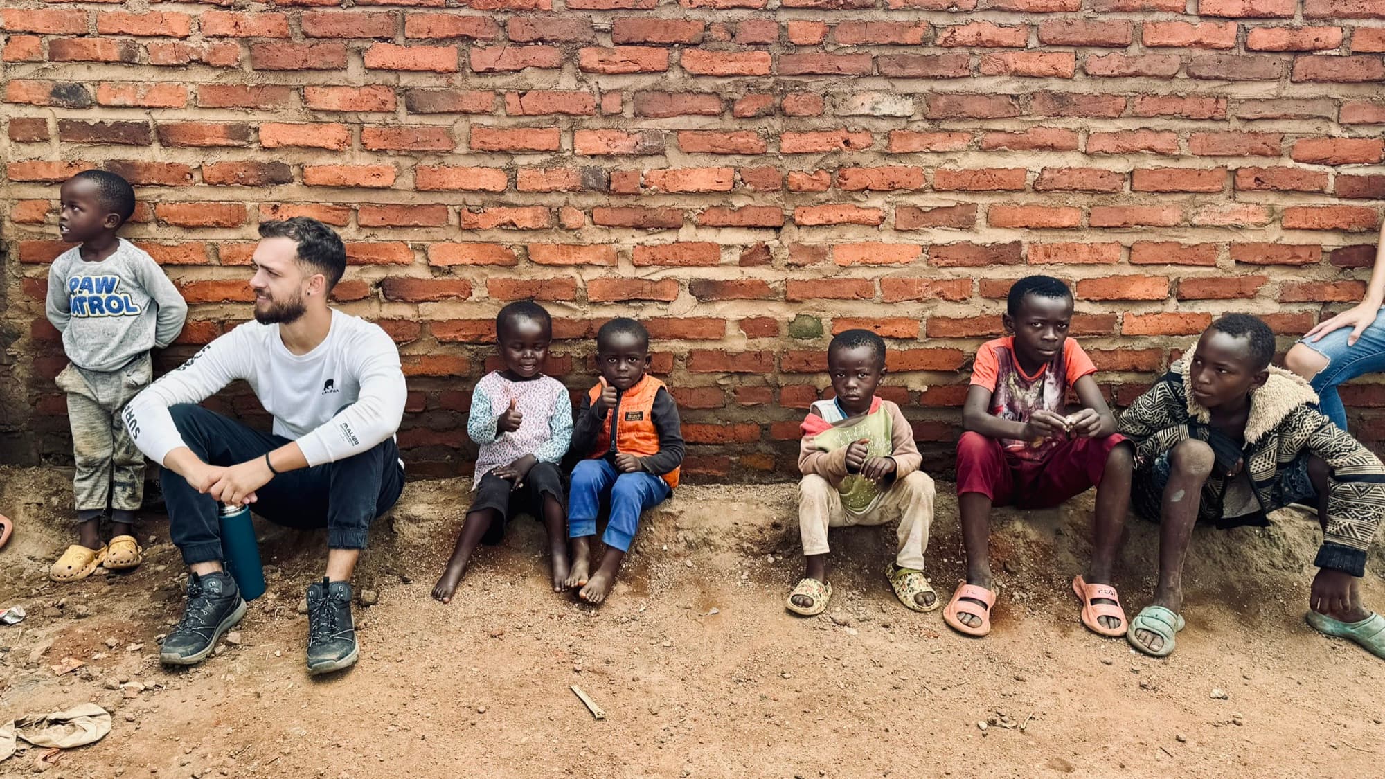 Felipe sentado no chão, encostado num muro de tijolos, ao lado de crianças do Malawi